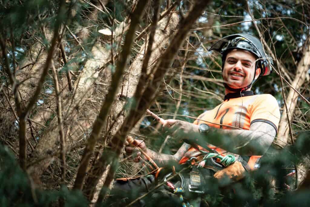 A person wearing an orange shirt, helmet, and safety gear smiles while positioned among tree branches, holding a rope and equipment—a typical scene for a tree surgeon in Edinburgh.