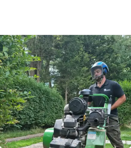 A tree surgeon in Edinburgh, wearing a helmet, operates a large green machine outdoors, surrounded by lush greenery and trees.